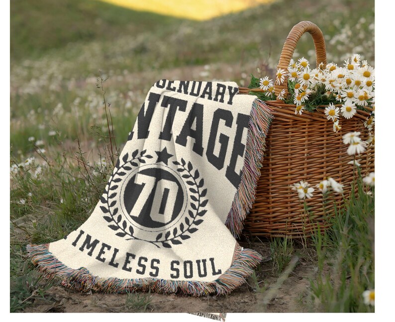 May include: A cream-colored blanket with black text reading "LEGENDARY VINTAGE 70 TIMELESS SOUL" is draped on the grass. A woven basket filled with daisies sits nearby. The blanket has a fringed edge and a star and laurel wreath design.