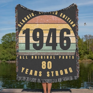 May include: A black woven blanket with a retro design. The blanket features the year "1946" in large numbers, with the text "CLASSIC. LIMITED EDITION" above and "ALL ORIGINAL PARTS 80 YEARS STRONG" below. The design includes a sunset-like color scheme.