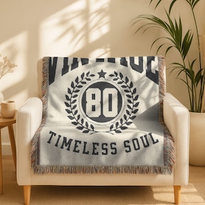 May include: Cream-colored throw blanket with black text and design. The blanket features the word "VINTAGE" at the top, a circular emblem with the number "80" inside a laurel wreath, and the words "TIMELESS SOUL" at the bottom. The blanket has a fringed edge.