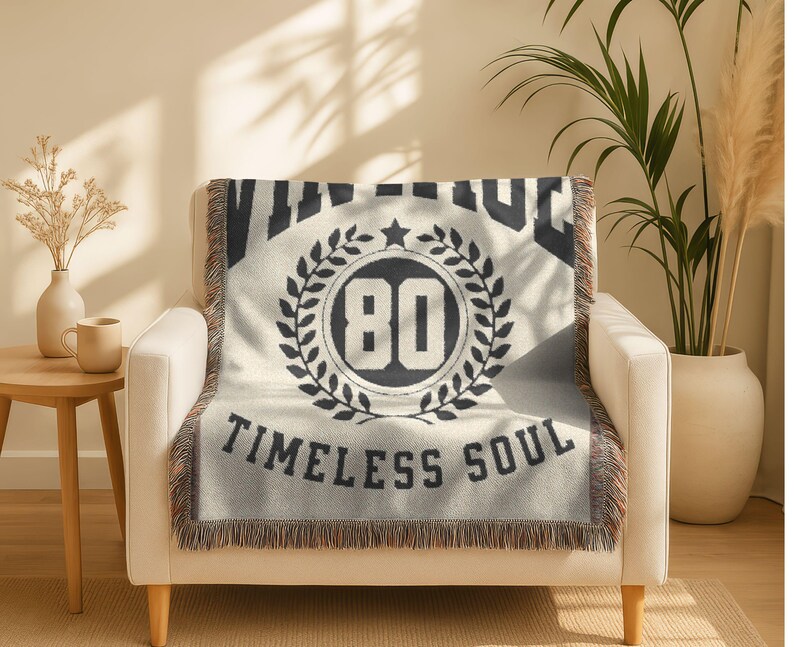 May include: Cream-colored throw blanket with black text and design. The blanket features the word "VINTAGE" at the top, a circular emblem with the number "80" inside a laurel wreath, and the words "TIMELESS SOUL" at the bottom. The blanket has a fringed edge.