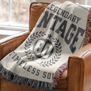 May include: A cream-colored throw blanket with the words "LEGENDARY VINTAGE" and "TIMELESS SOUL" printed in dark gray. The blanket features a circular design with the number "80" inside a laurel wreath. The blanket is draped over a brown leather armchair.
