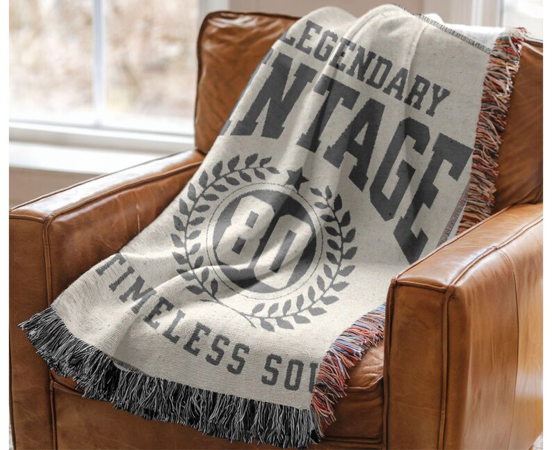 May include: A cream-colored throw blanket with the words "LEGENDARY VINTAGE" and "TIMELESS SOUL" printed in dark gray. The blanket features a circular design with the number "80" inside a laurel wreath. The blanket is draped over a brown leather armchair.
