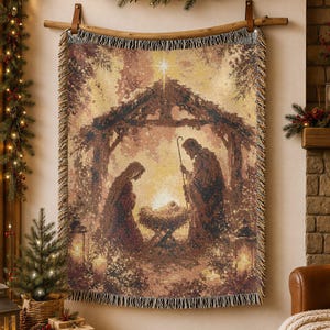 May include: A woven tapestry throw blanket featuring a nativity scene with a central manger, figures of Mary and Joseph, and a bright star above. The blanket has a fringed edge and is displayed hanging from a wooden rod with leather straps.