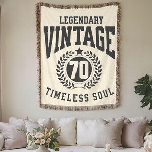 May include: Cream-colored woven throw blanket with black text that reads "LEGENDARY VINTAGE 70 TIMELESS SOUL." The number 70 is inside a laurel wreath. The blanket has a fringed edge and is hanging on a wall above a white sofa.