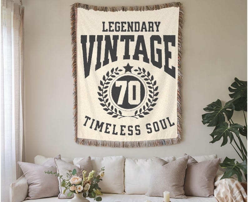 May include: Cream-colored woven throw blanket with black text that reads "LEGENDARY VINTAGE 70 TIMELESS SOUL." The number 70 is inside a laurel wreath. The blanket has a fringed edge and is hanging on a wall above a white sofa.