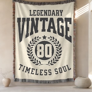 May include: Cream-colored throw blanket with black text that reads "LEGENDARY VINTAGE TIMLESS SOUL". The word "VINTAGE" is above a circular design with the number "80" inside. The blanket has a fringed edge.