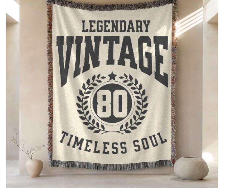 May include: Cream-colored throw blanket with black text that reads "LEGENDARY VINTAGE TIMLESS SOUL". The word "VINTAGE" is above a circular design with the number "80" inside. The blanket has a fringed edge.