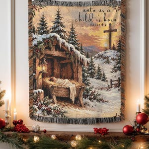 May include: A woven tapestry throw blanket featuring a winter nativity scene with a manger, a glowing lantern, snow-covered pine trees, and a cross on a hill. The text reads: unto us a child is born, Isaiah 9:6. The blanket has a fringed edge.