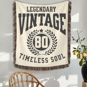 May include: Cream-colored woven throw blanket with the words "LEGENDARY VINTAGE TIMELESS SOUL" in black. The number "80" is inside a laurel wreath. The blanket has a fringed edge and is hanging on a wall.