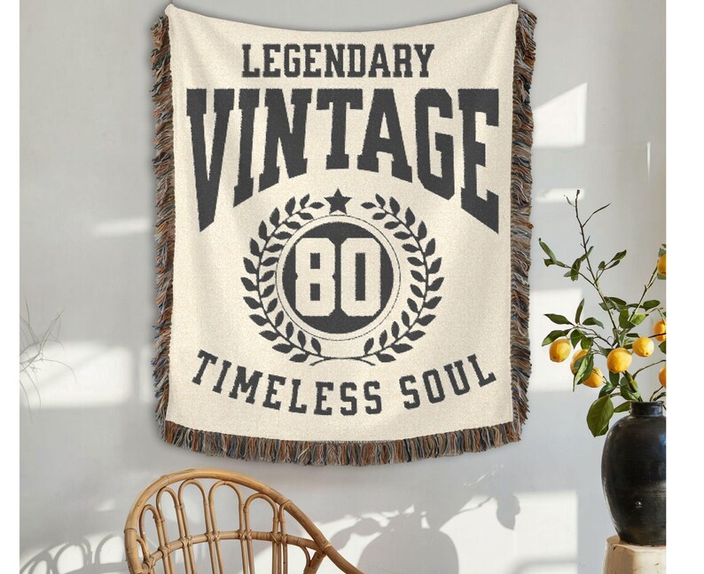 May include: Cream-colored woven throw blanket with the words "LEGENDARY VINTAGE TIMELESS SOUL" in black. The number "80" is inside a laurel wreath. The blanket has a fringed edge and is hanging on a wall.