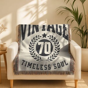 May include: Cream-colored throw blanket with black text and design. The blanket features the word "VINTAGE" above a circular emblem with the number "70" and the words "TIMELESS SOUL". The blanket is draped over a white armchair.