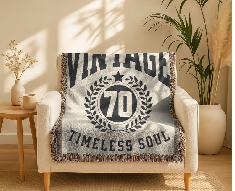 May include: Cream-colored throw blanket with black text and design. The blanket features the word "VINTAGE" above a circular emblem with the number "70" and the words "TIMELESS SOUL". The blanket is draped over a white armchair.