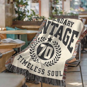 May include: Cream-colored woven throw blanket with black text that reads "LEGENDARY VINTAGE" and "TIMELESS SOUL." A circular emblem features the number "70" inside a laurel wreath. The blanket has a fringed edge with red, black, and white threads.