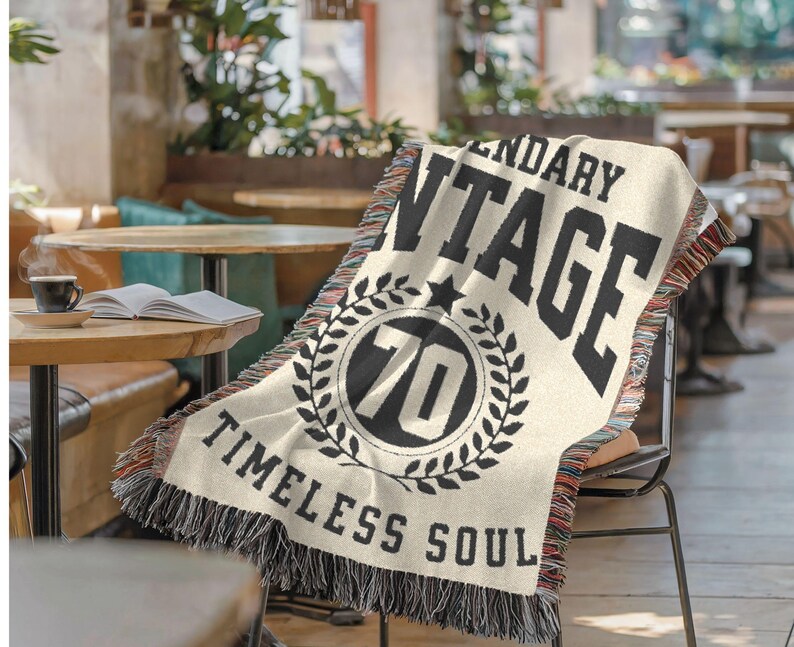 May include: Cream-colored woven throw blanket with black text that reads "LEGENDARY VINTAGE" and "TIMELESS SOUL." A circular emblem features the number "70" inside a laurel wreath. The blanket has a fringed edge with red, black, and white threads.