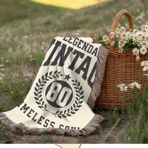 May include: A cream-colored throw blanket with black text reading "LEGENDARY VINTAGE 80 TIMELESS SOUL" is draped on the grass. A woven basket filled with daisies sits nearby. The blanket has a fringed edge.
