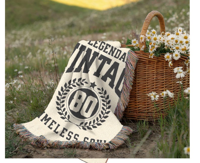 May include: A cream-colored throw blanket with black text reading "LEGENDARY VINTAGE 80 TIMELESS SOUL" is draped on the grass. A woven basket filled with daisies sits nearby. The blanket has a fringed edge.