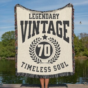 May include: Cream-colored woven blanket with black text that reads "LEGENDARY VINTAGE 70 TIMELESS SOUL." The blanket has a fringed edge with a mix of colors. The number 70 is inside a circular wreath design.