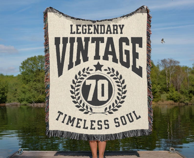May include: Cream-colored woven blanket with black text that reads "LEGENDARY VINTAGE 70 TIMELESS SOUL." The blanket has a fringed edge with a mix of colors. The number 70 is inside a circular wreath design.