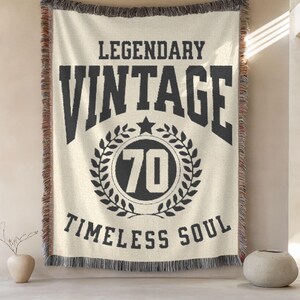 May include: Cream-colored woven throw blanket with black text that reads "LEGENDARY VINTAGE 70 TIMELESS SOUL." The blanket has a black and brown fringe.