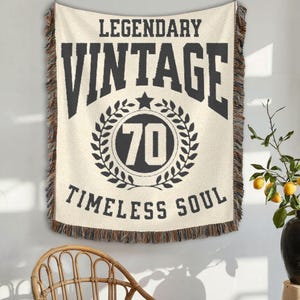 May include: Cream-colored woven throw blanket with black text that reads "LEGENDARY VINTAGE 70 TIMELESS SOUL." The blanket has a fringed edge and is hanging on a wall. A wicker chair is in the foreground.