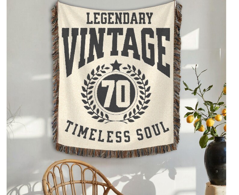 May include: Cream-colored woven throw blanket with black text that reads "LEGENDARY VINTAGE 70 TIMELESS SOUL." The blanket has a fringed edge and is hanging on a wall. A wicker chair is in the foreground.