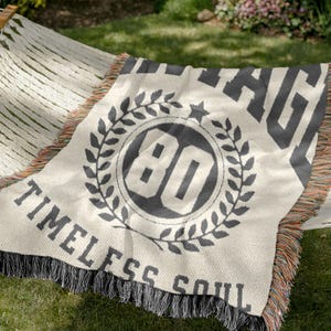 May include: Cream-colored throw blanket with black text and design. The blanket features the number "80" inside a laurel wreath, with the words "VINTAGE" and "TIMELESS SOUL". The blanket has a fringed edge.