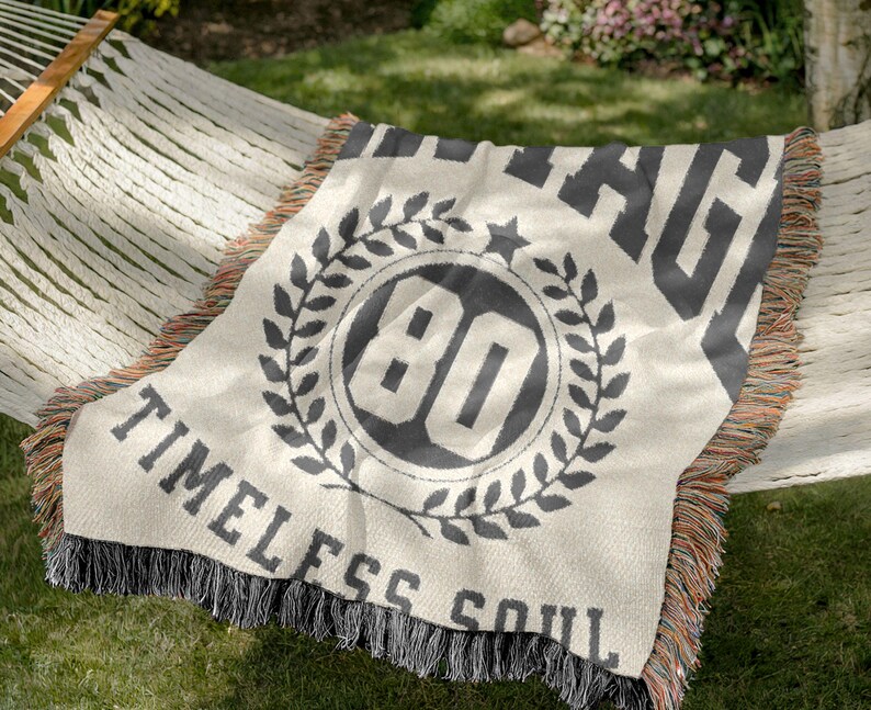 May include: Cream-colored throw blanket with black text and design. The blanket features the number "80" inside a laurel wreath, with the words "VINTAGE" and "TIMELESS SOUL". The blanket has a fringed edge.