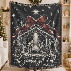 May include: A black woven throw blanket featuring a nativity scene with a large red bow at the top. The design includes a bright star, figures around a manger, and the text "the greatest gift of all" followed by "Matthew 1:21" in white script.