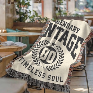 May include: Cream-colored woven throw blanket with black text that reads "LEGENDARY VINTAGE" and "TIMELESS SOUL." The blanket features a circular design with the number "80" inside a laurel wreath. The edges have a fringed trim.