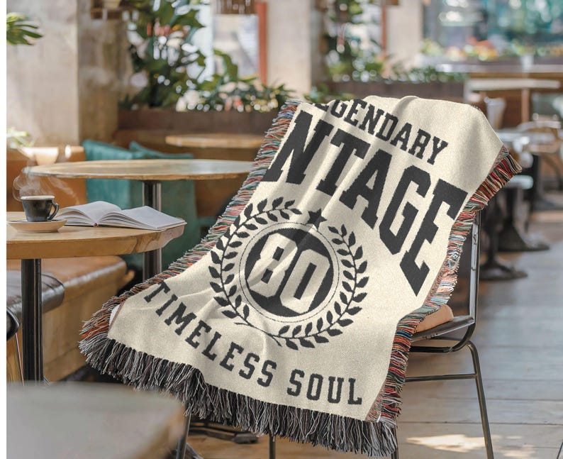 May include: Cream-colored woven throw blanket with black text that reads "LEGENDARY VINTAGE" and "TIMELESS SOUL." The blanket features a circular design with the number "80" inside a laurel wreath. The edges have a fringed trim.
