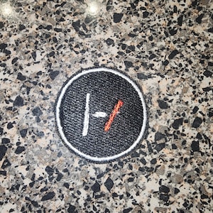 May include: A circular embroidered patch featuring a black background with a white stitched border. In the center, a white vertical line is positioned next to a red diagonal line, forming a stylized symbol on the textured black fabric surface.
