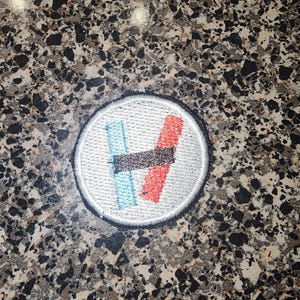 May include: A circular embroidered patch featuring a white background with a black stitched border. The design consists of a vertical light blue rectangle, a vertical red rectangle, and a horizontal black rectangle layered across the center.