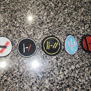 May include: Five circular and oval embroidered patches featuring various graphic designs. The first is white with blue and red lines, the second is black with white and red lines, the third is black with yellow lines, the fourth is light blue with a pink trident, and the fifth is red with black lines.