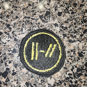 May include: A circular embroidered patch featuring a black background with a yellow stitched border and a yellow stitched logo in the center. The logo consists of two vertical parallel lines followed by a hyphen and two slanted parallel lines.