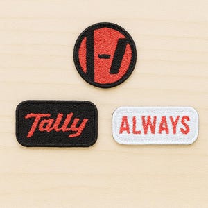 Puede incluir: Tres parches bordados sobre una superficie de madera clara. El parche superior es un círculo rojo y negro con un diseño estilizado. Debajo hay dos parches rectangulares: uno negro con "Tally" en rojo y otro blanco con "ALWAYS" en rojo.