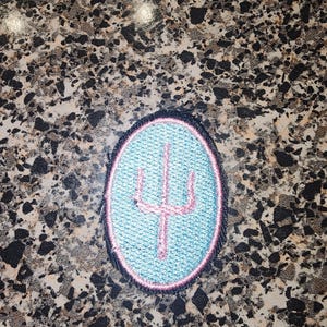 May include: An oval-shaped embroidered patch featuring a light blue background with a pink trident symbol in the center. The patch is outlined with a thin pink border and a thicker black outer edge, creating a distinct contrast in the design.