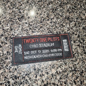 May include: A rectangular black faux leather patch embroidered to resemble a concert ticket. It features red and white stitching with the text TWENTY ONE PILOTS, OHIO STADIUM, SAT OCT 17 2026 8:00 PM, WITH DEATH CAB FOR CUTIE, and ONE ADMIT.