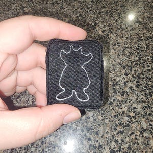 Pode incluir: Um emblema retangular bordado em preto com o contorno branco costurado de uma criatura estilizada semelhante a um urso. O emblema tem uma borda preta espessa e uma superfície de tecido texturizado, exibindo um design gráfico simples.