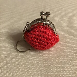 Può includere: Un piccolo portamonete rotondo all'uncinetto rosso, con una chiusura in metallo color argento e un portachiavi. La borsetta ha un aspetto strutturato grazie alle maglie all'uncinetto. La chiusura presenta due piccoli accenti decorativi a forma di sfera.