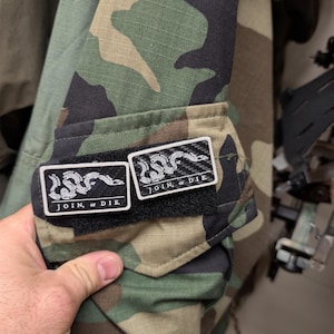 May include: A camouflage patterned jacket sleeve featuring two rectangular black and white patches. Each patch displays a graphic of a segmented snake and the text JOIN, or DIE. The patches are attached to a hook-and-loop fastener strip.