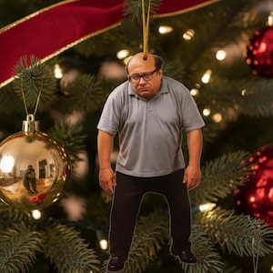 May include: A Christmas tree ornament featuring a person wearing glasses, a gray polo shirt, and black pants. The ornament is hanging from a gold string against a backdrop of a decorated Christmas tree with red and gold ornaments.
