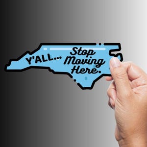 May include: A light blue sticker in the shape of North Carolina with the text "Y'all... Stop Moving Here." The sticker has a black outline and is held by a hand against a gradient background.