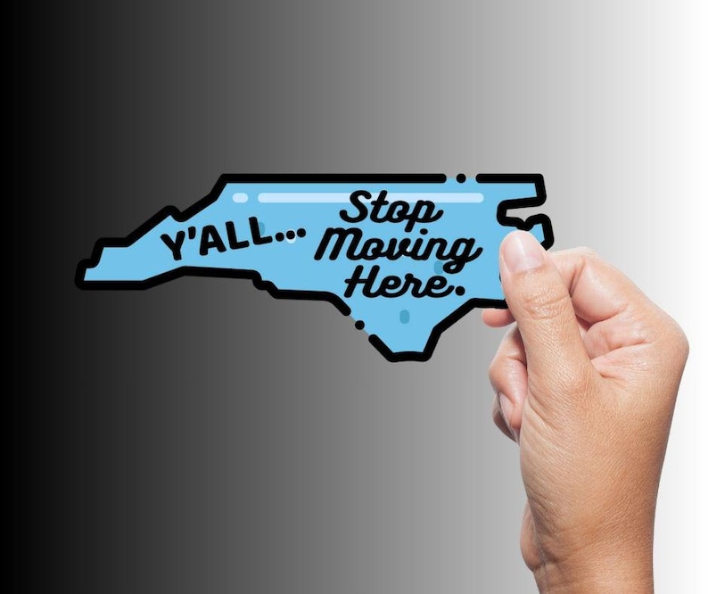 May include: A light blue sticker in the shape of North Carolina with the text "Y'all... Stop Moving Here." The sticker has a black outline and is held by a hand against a gradient background.