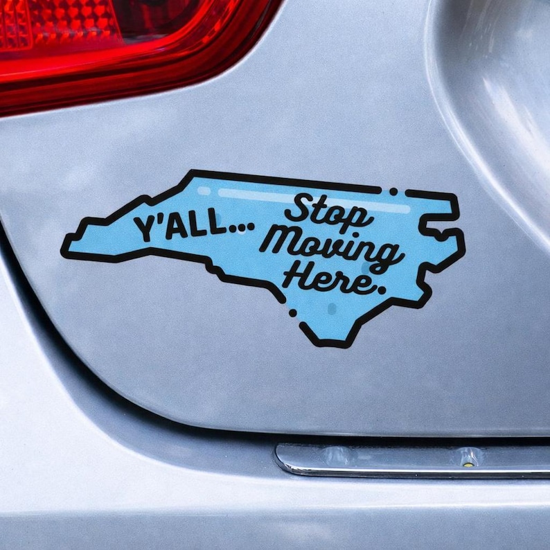 May include: A blue sticker in the shape of North Carolina with the text "Y'ALL... Stop Moving Here." The sticker has a black outline and is affixed to a silver car.