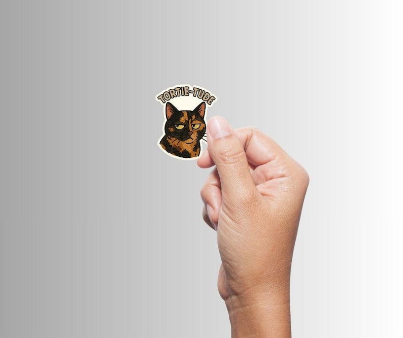 May include: A sticker featuring a tortoiseshell cat with a grumpy expression. The sticker has the word "TORTIE-TUDE" in a circular design above the cat's head. The cat's fur is a mix of black, brown, and orange.