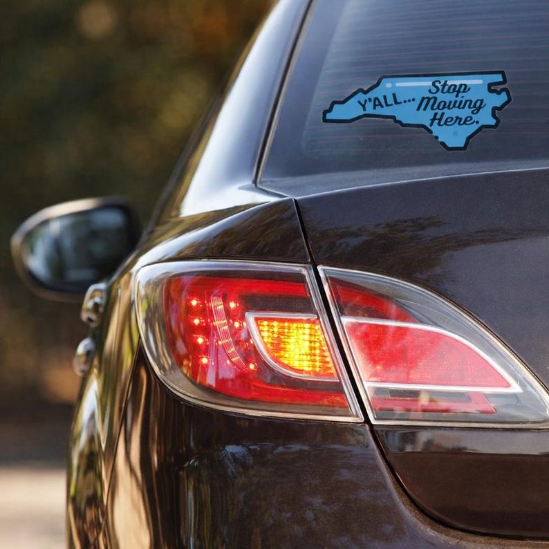 May include: A car's rear window features a blue sticker shaped like North Carolina. The sticker says "Y'all... Stop Moving Here" with a light blue outline. The car's taillight is red and orange.