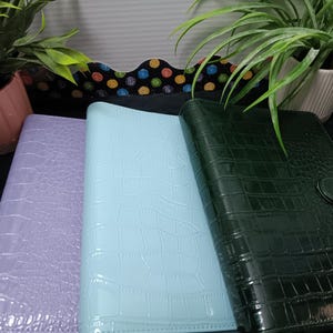 May include: Three faux-leather binders in pastel colors: lavender, light blue, and dark green. Each binder has a crocodile skin texture and a snap closure. The binders are displayed with potted plants in the background.