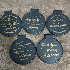以下が含まれることがあります： 金色の文字が入った、濃い青色の丸いコンパクトミラー5個セット。各ミラーには、「You Rock!」や「You're Awesome」など、異なるインスピレーションを与えるメッセージが書かれています。ミラーの上部には小さなハンドルが付いています。