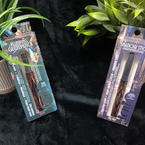 May include: Two cosmetic products in packaging are displayed. One is a "Plumping Gloss Stick" in a teal box with the word "Rose". The other is a "Shadow Stick" in a black box. Both products are in clear plastic packaging.