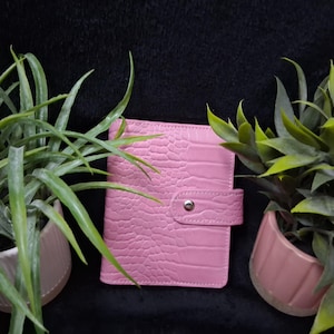 May include: A rectangular pink notebook with a textured crocodile skin pattern on the cover. The notebook features a central vertical strap that secures with a single silver-toned metal snap button closure, positioned between two potted green plants.